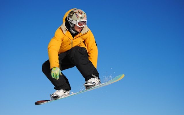 Snowboardfahrer in gelber Jacke springt über eine Schneepiste unter klarem blauen Himmel.