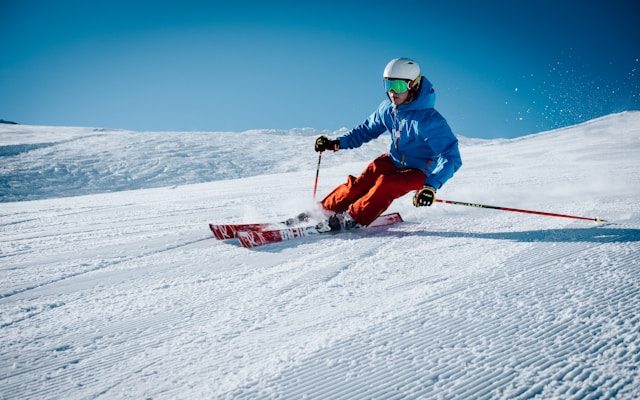 Skifahrer in blauer Jacke und roten Hosen, der auf einer schneebedeckten Piste mit Schwung fährt.