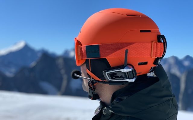 Oranger Skihelm mit integrierter Kommunikationsausrüstung, getragen von einer Person in den Bergen.